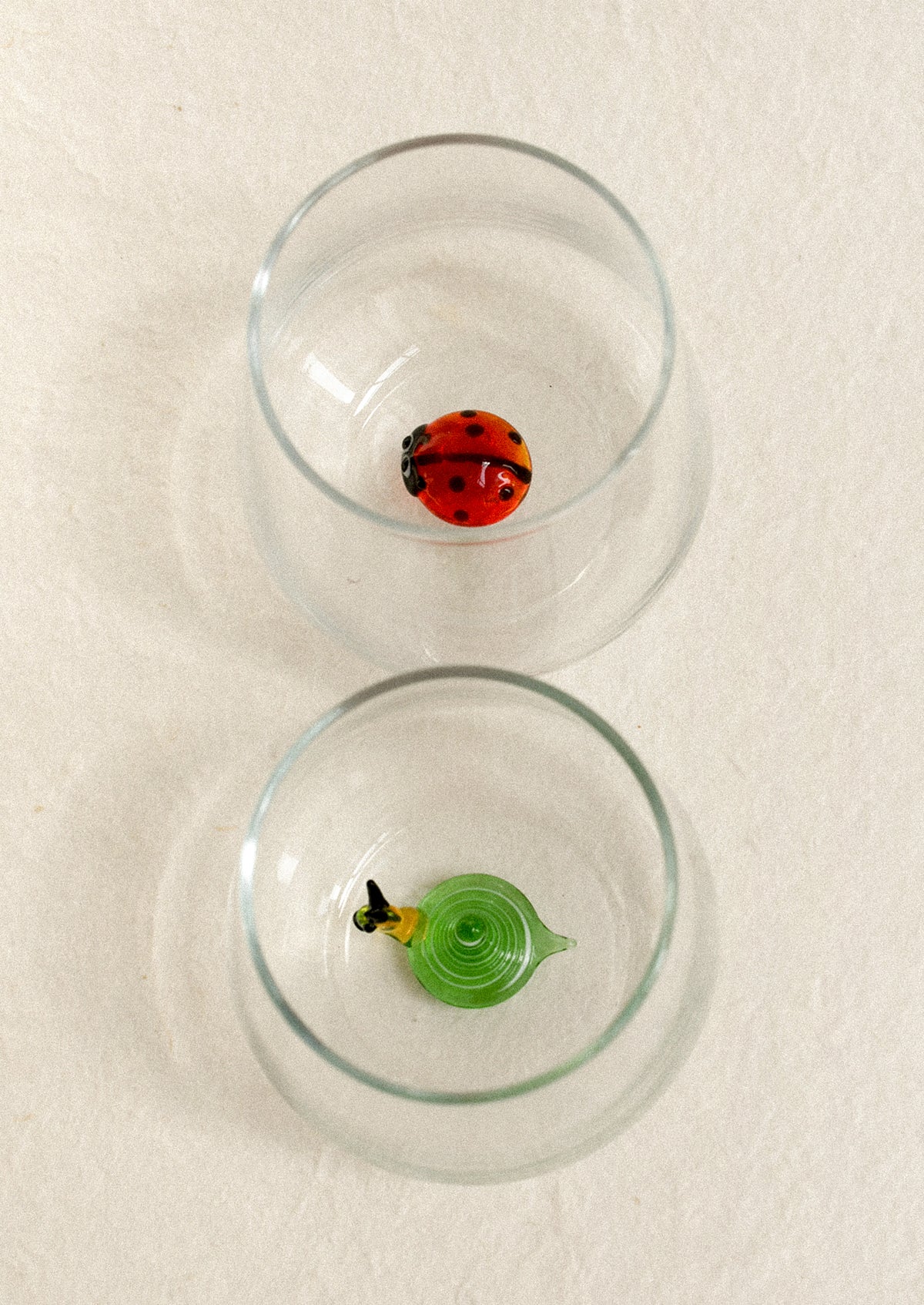 Two clear glasses, each containing a small decorative glass figure: a red ladybug in the top glass and a green snail form in the bottom style.