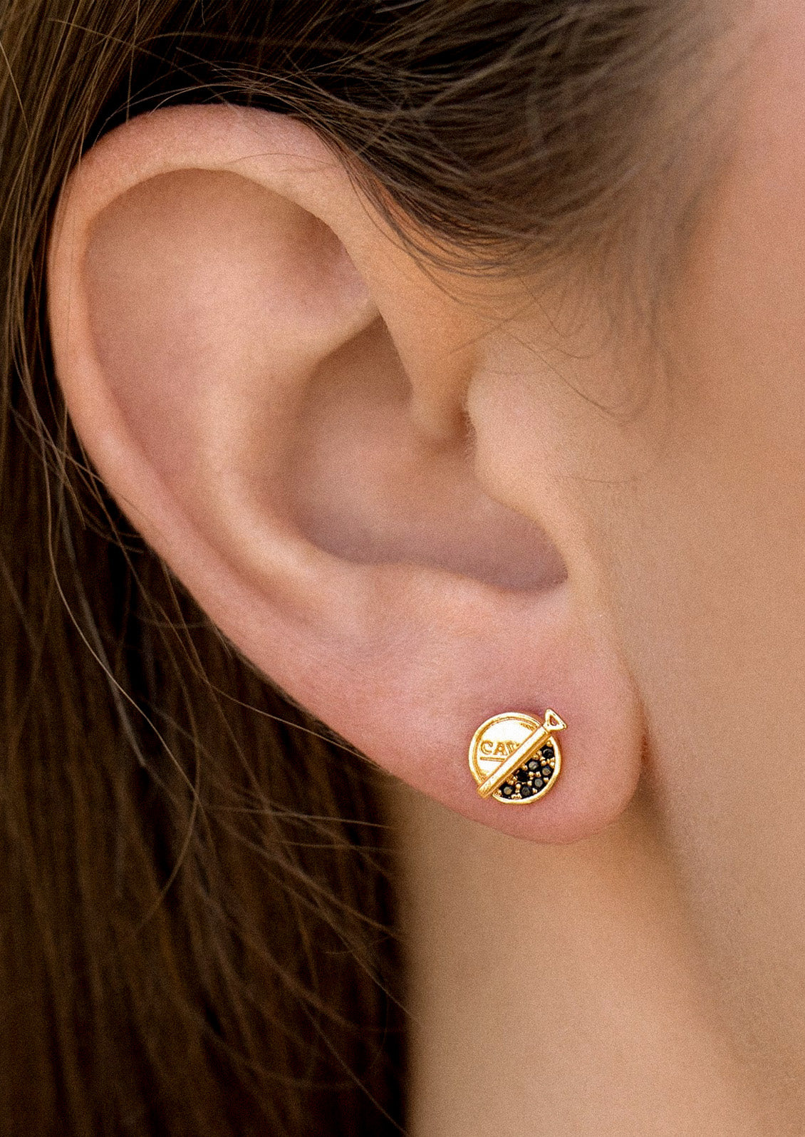 A woman wearing a gold plated stud earring in the shape of a caviar can with black and gold gems imitating caviar.