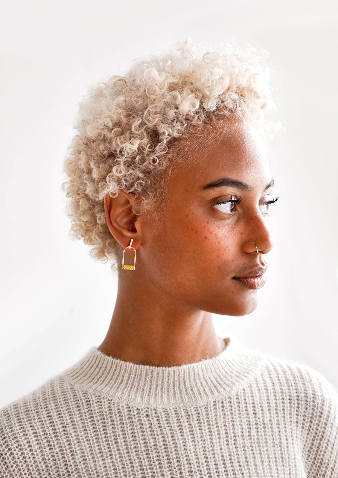Model wears arced gold earrings with yellow beads and a cream colored sweater.