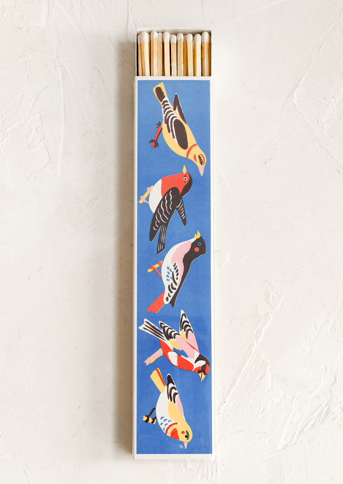 A long rectangular matchbox with a blue background printed with stylized birds in yellow, red, black, white, and pink, shown with matches sticking out at the top against a light textured wall.