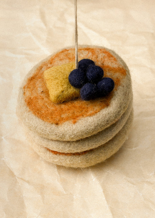 A felted wool miniature stack of three pancakes topped with orange-brown syrup, a yellow butter piece, and dark blue felted blueberries, with a metal pin sticking up from the center, set on a beige textured paper background.