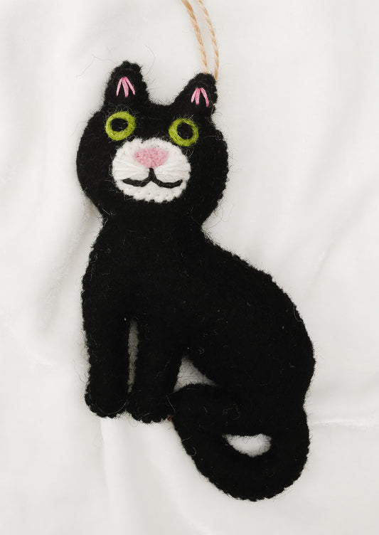 A black felt cat ornament with embroidered green eyes, pink nose and ear accents, and a white chin, hanging from a tan string against a white fabric background.