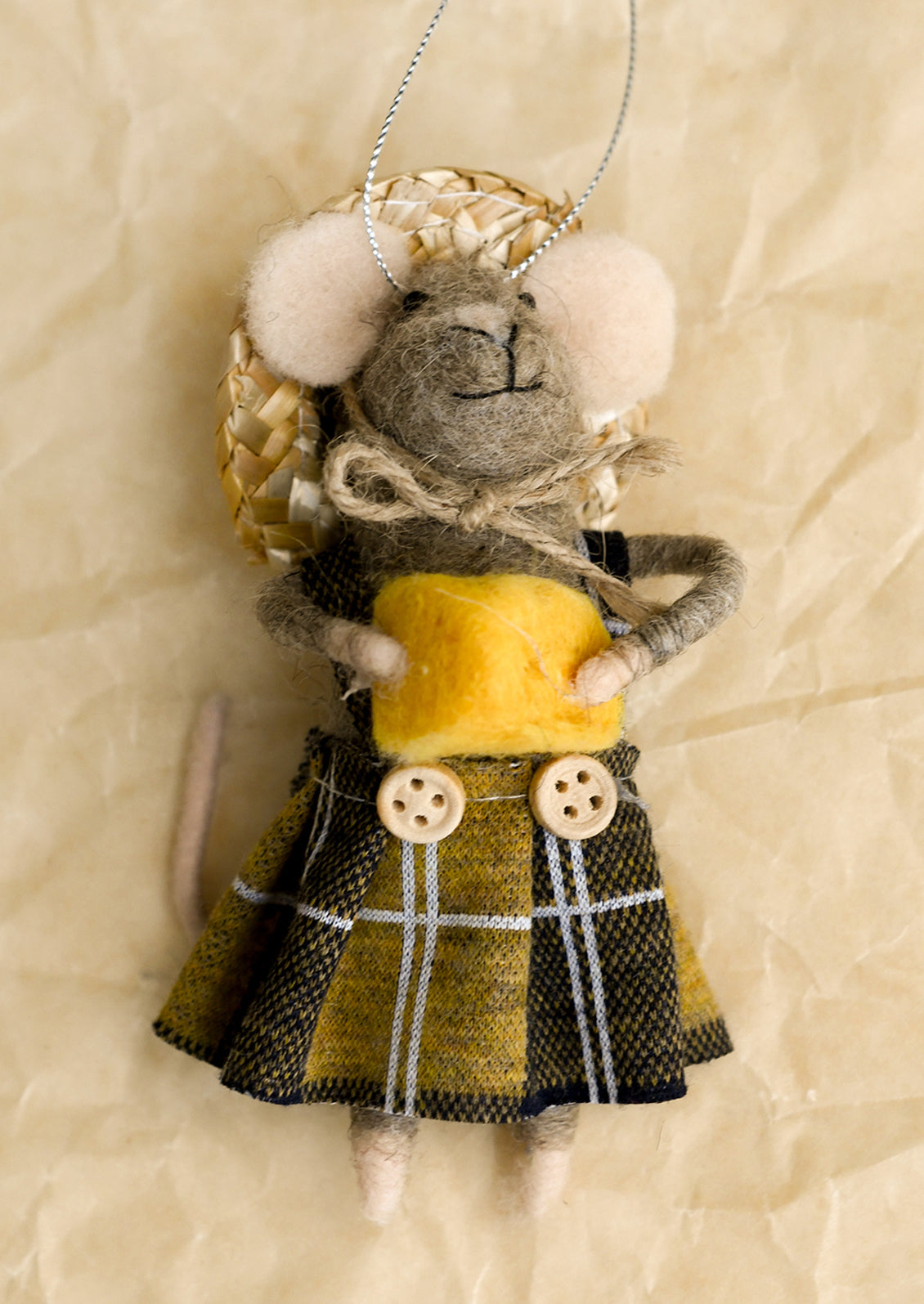 A handmade felted wool mouse ornament dressed in a yellow shirt and a black and yellow plaid skirt, wearing a woven straw hat with white felted wool ears, hanging against a textured beige background.