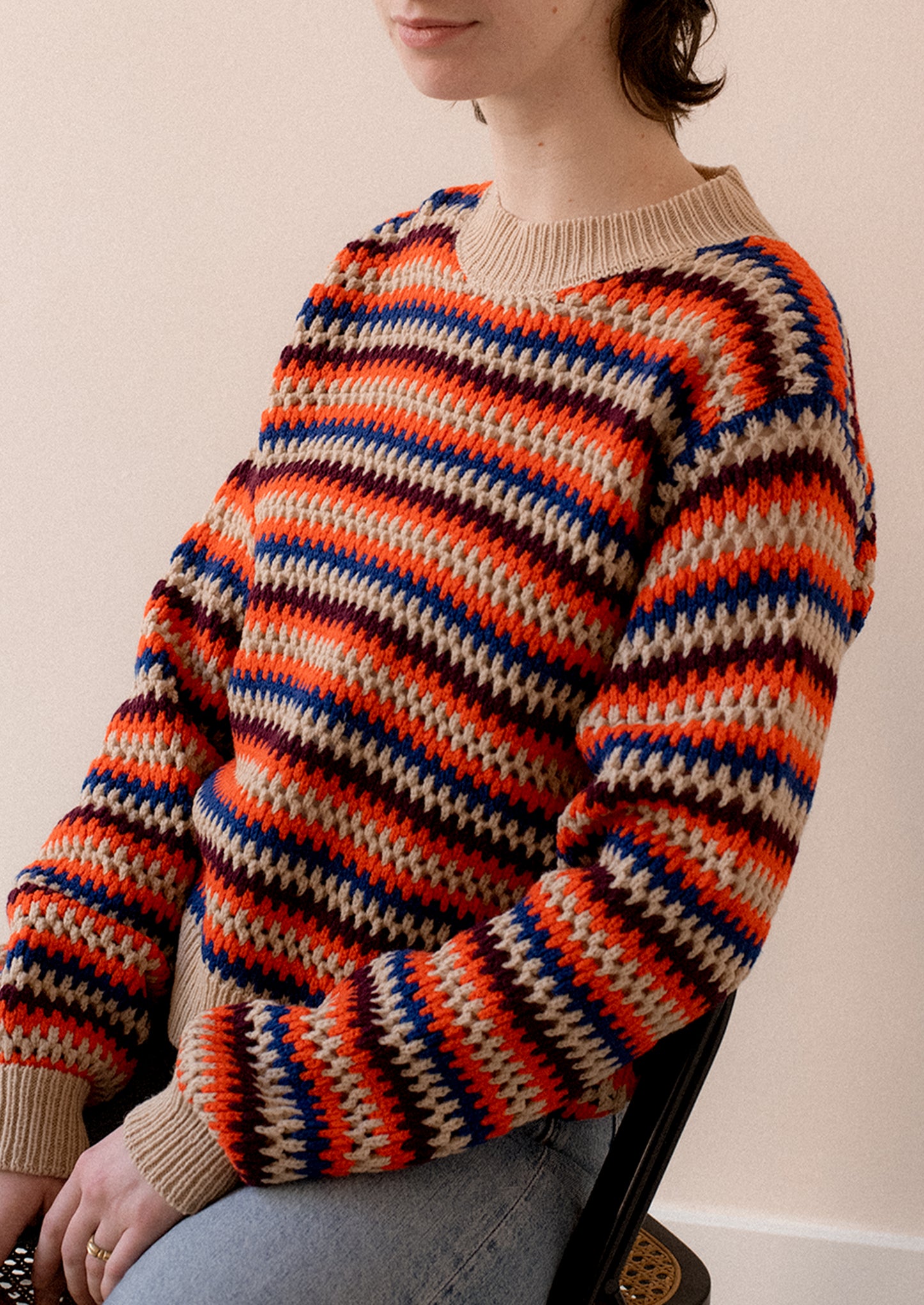 A woman wearing a crochet knit striped sweater in cream with orange, maroon and blue stripes.