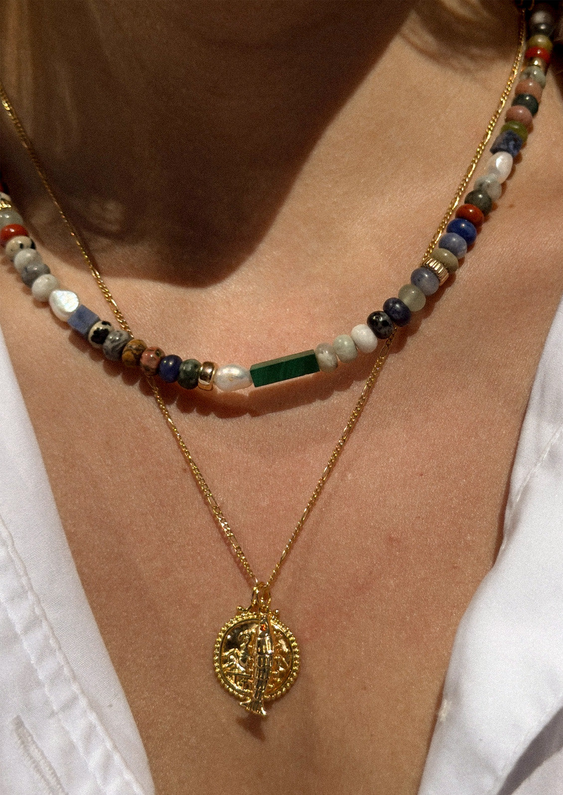 A woman wearing a gold carabiner necklace adorned with various colored gemstones in muted tones along with two other gold necklaces.