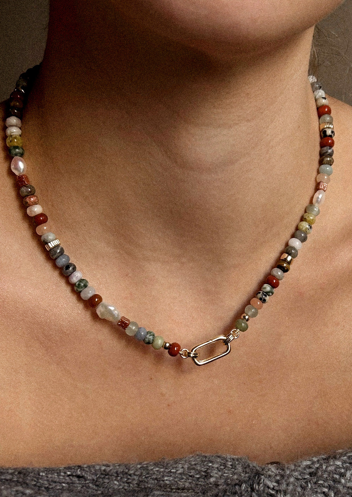 A woman wearing a gold carabiner necklace adorned with various colored gemstones in muted tones.