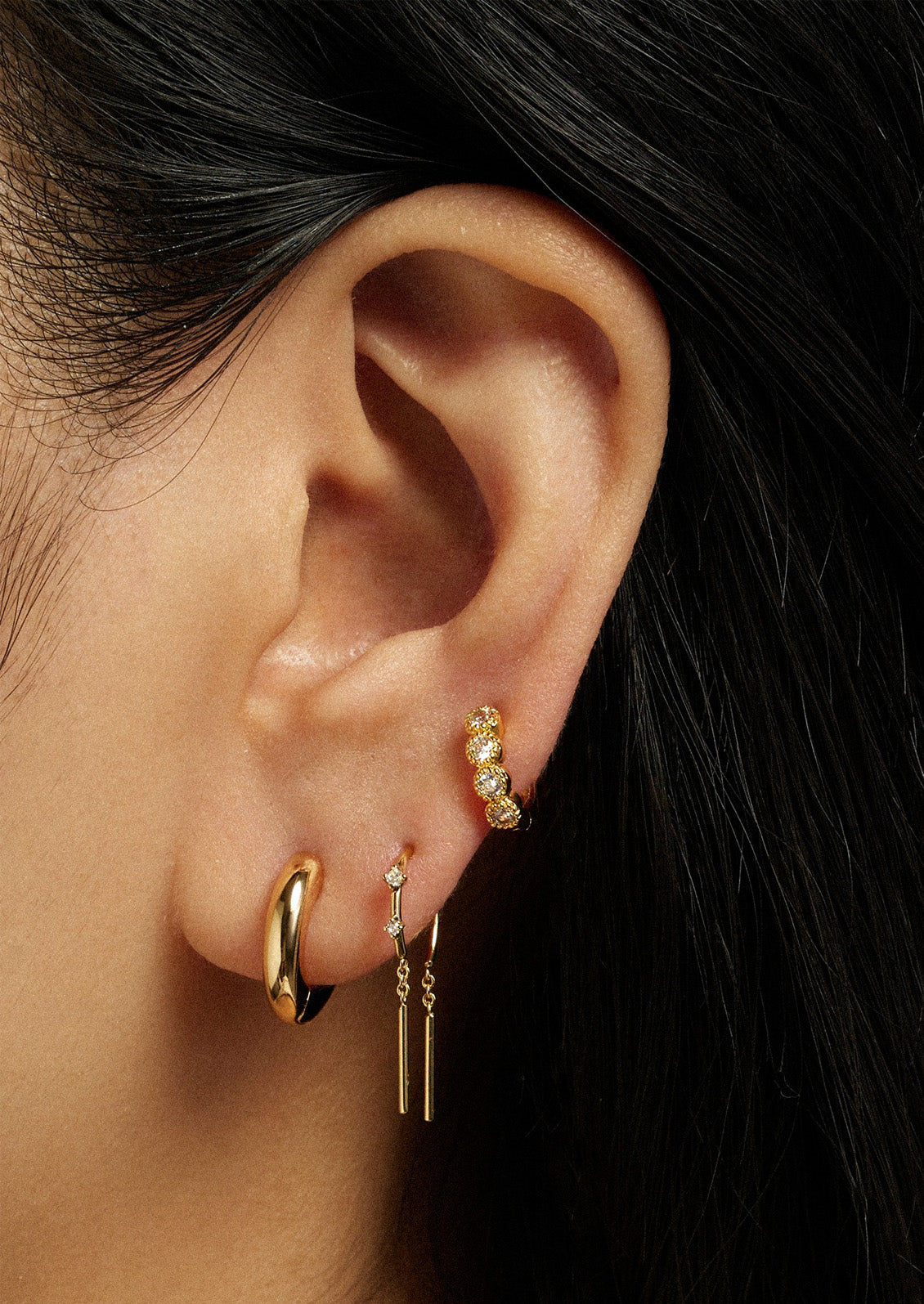 A woman wearing three different gold earrings on her ear including two hoop earrings and one threader earring.