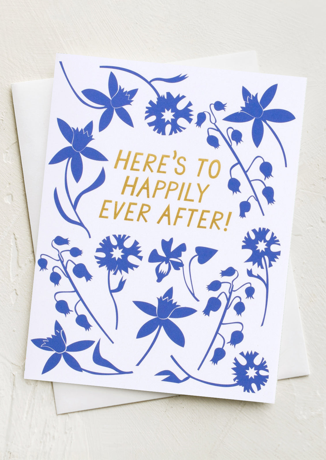 A greeting card with cobalt blue floral pattern, text reads "Here's to Happily Ever After!".