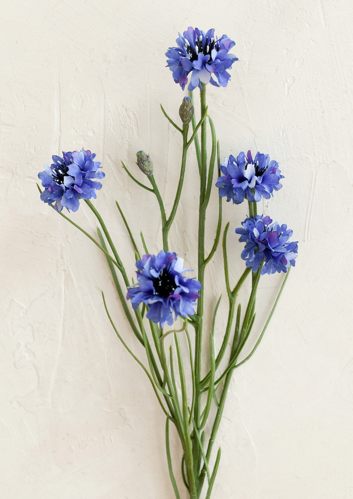 Artificial blue cornflowers with green stems and buds, arranged against a textured off-white background.