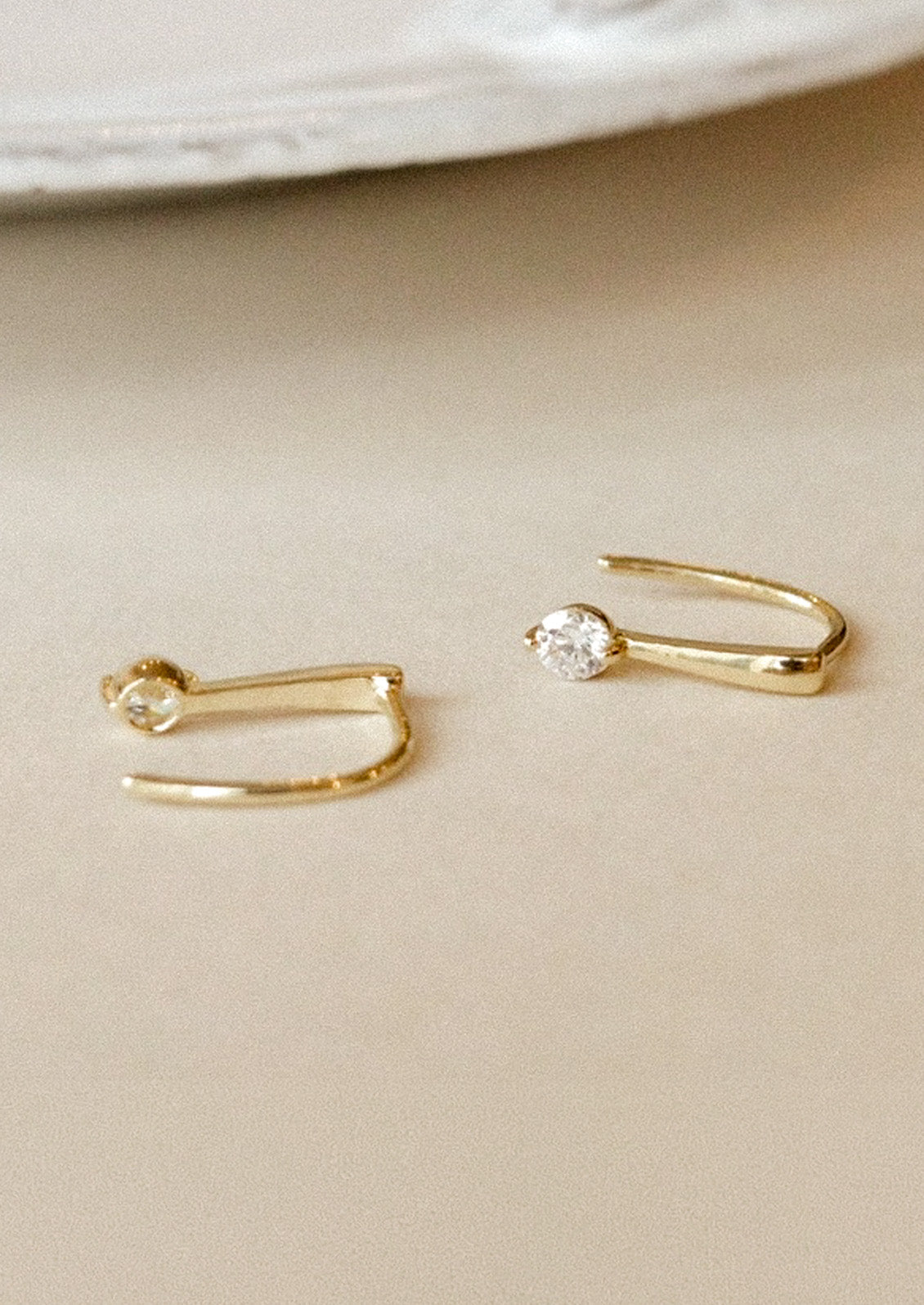 A pair of gold hook earrings with a cubic zirconia solitaire gem laying on a beige surface.