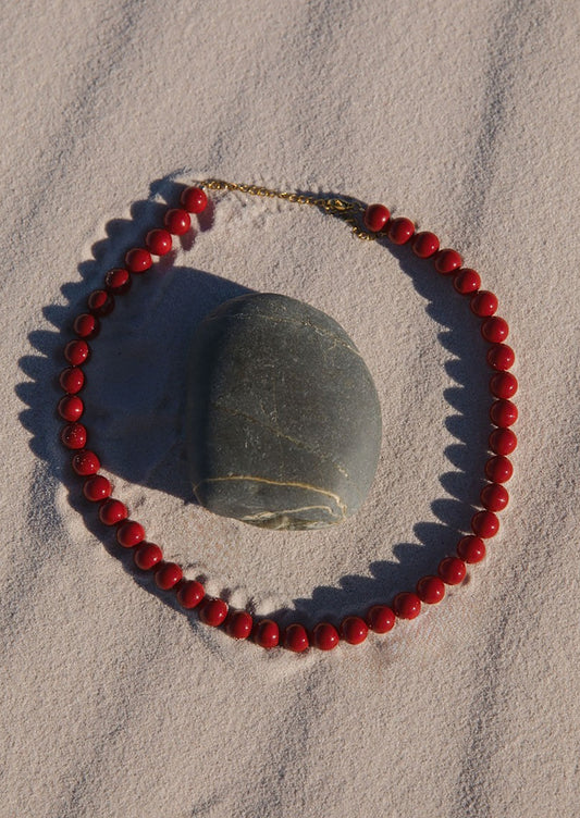 Red beaded necklace on a sandy surface with a stone in the background