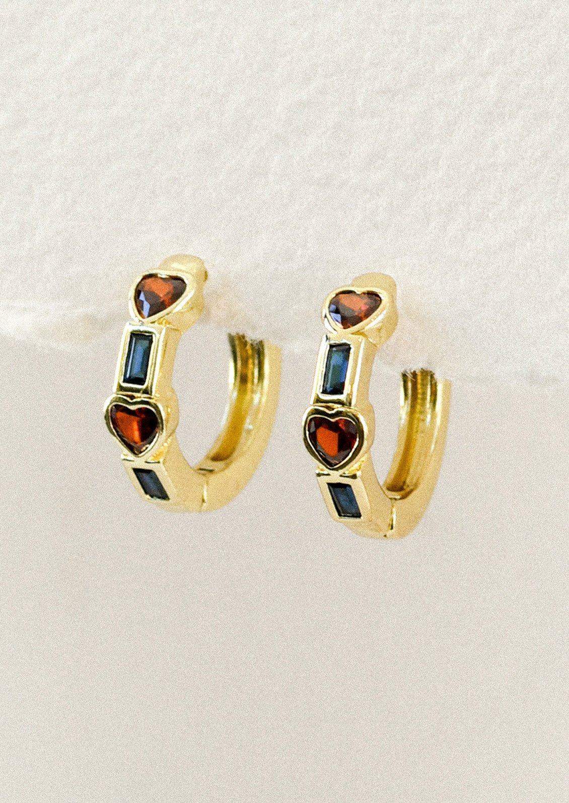 A pair of gold huggie hoop earrings with alternating red heart and blue rectangular inlaid gems.