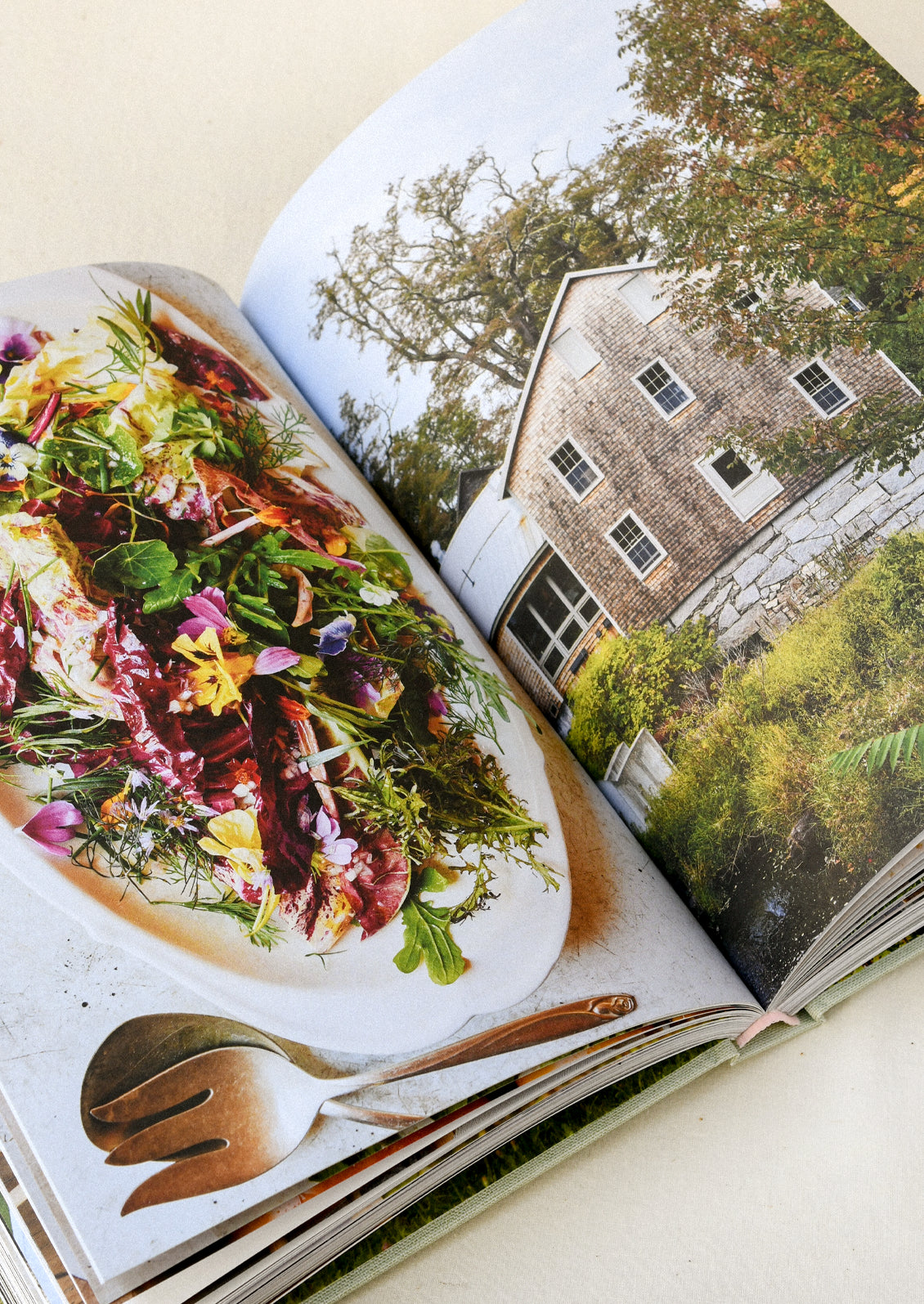 A cookbook open on a table showing a salad on one page and an old country house on the other.