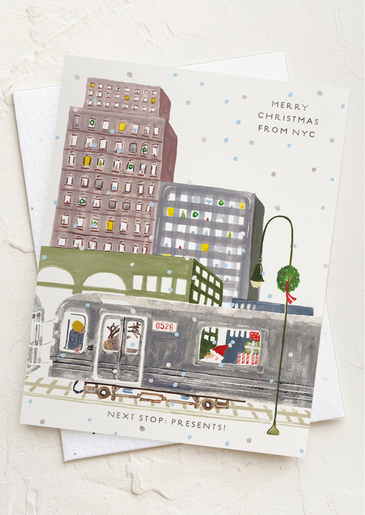 A Christmas-themed greeting card featuring an illustrated city street scene with a subway train, buildings, and a lit lamp post, shown on a textured white background with a white envelope behind it.