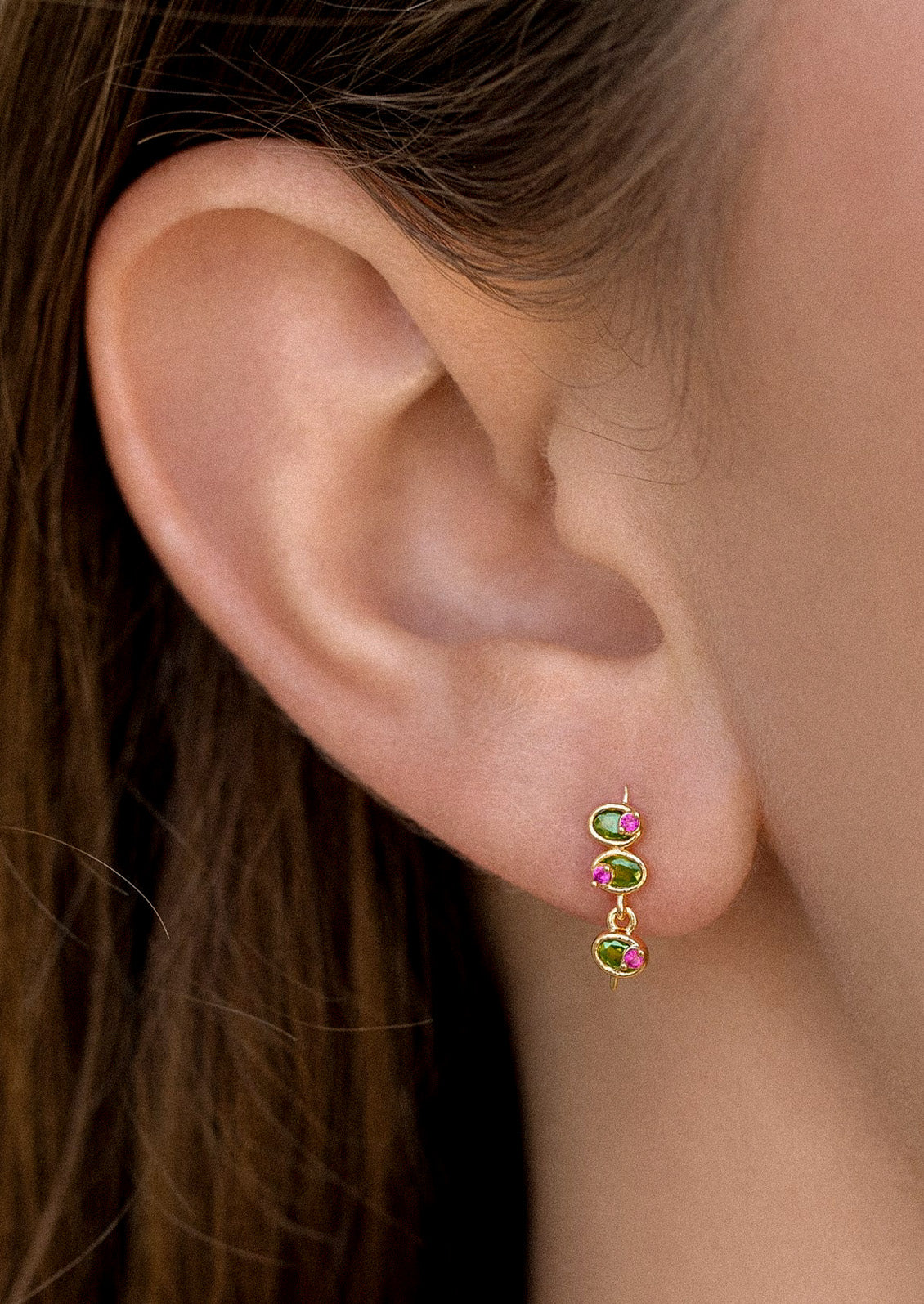 A woman wearing a long gold stud earring in the shape of olives with green and pink gems.