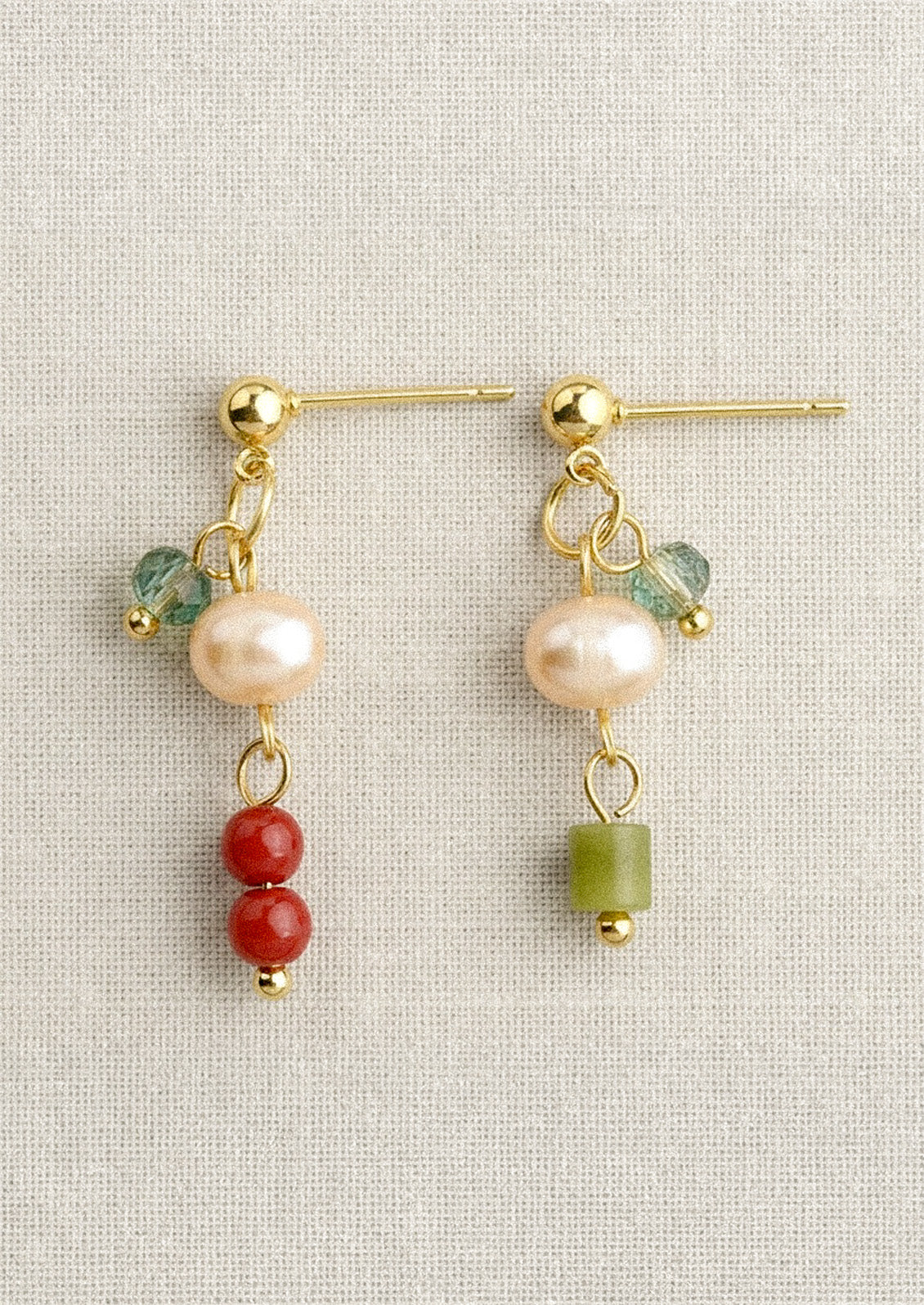 A pair of gold dangling earrings each with a freshwater pearl and differing blue, green, and red gems on a linen fabric.