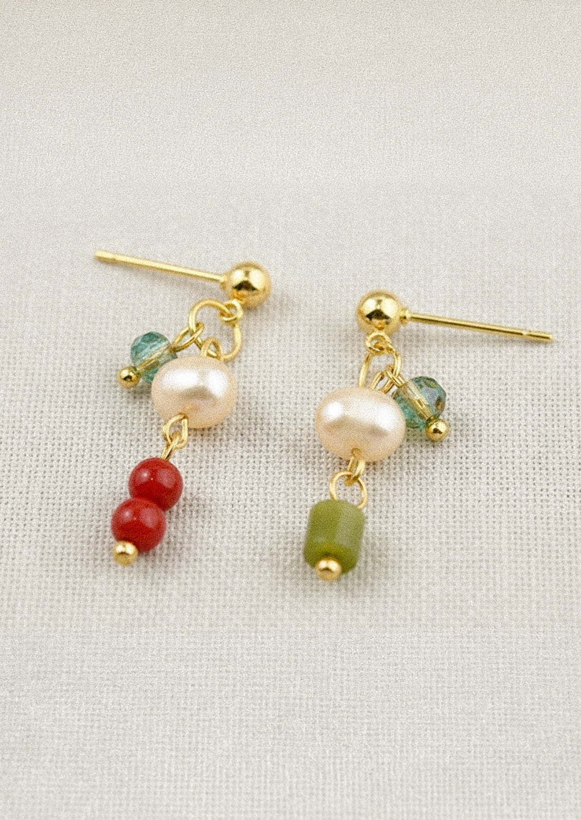 A pair of gold dangling earrings each with a freshwater pearl and differing blue, green, and red gems on a linen fabric.