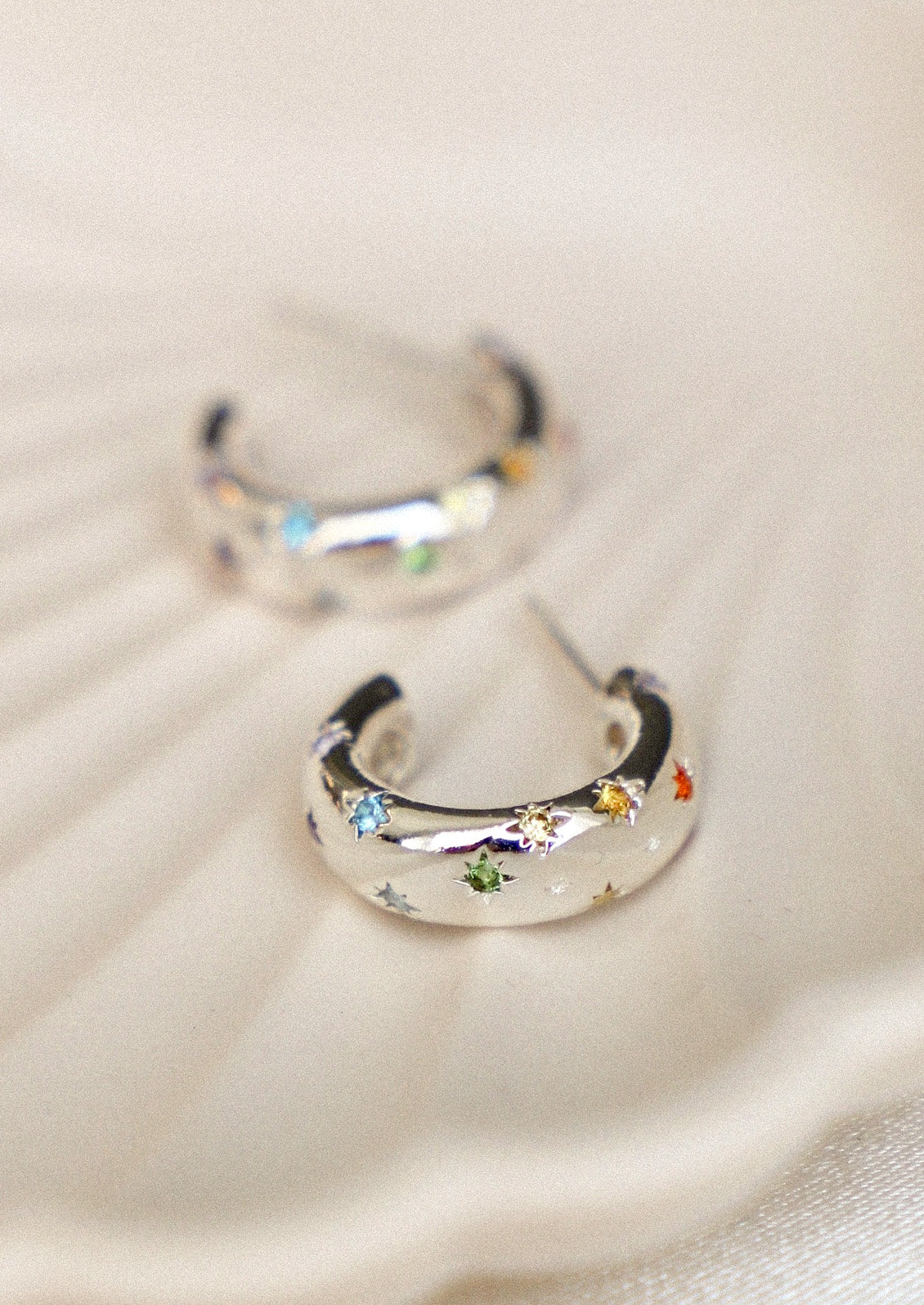 A pair of silver crescent-shaped hoop earrings decorated with small multicolored star and flower charms, resting on a textured beige fabric background.