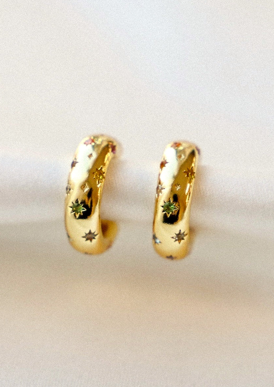 A pair of gold hoop earrings with small starbursts throughout adorned with various colored gems in the centers attached to a white fabric.