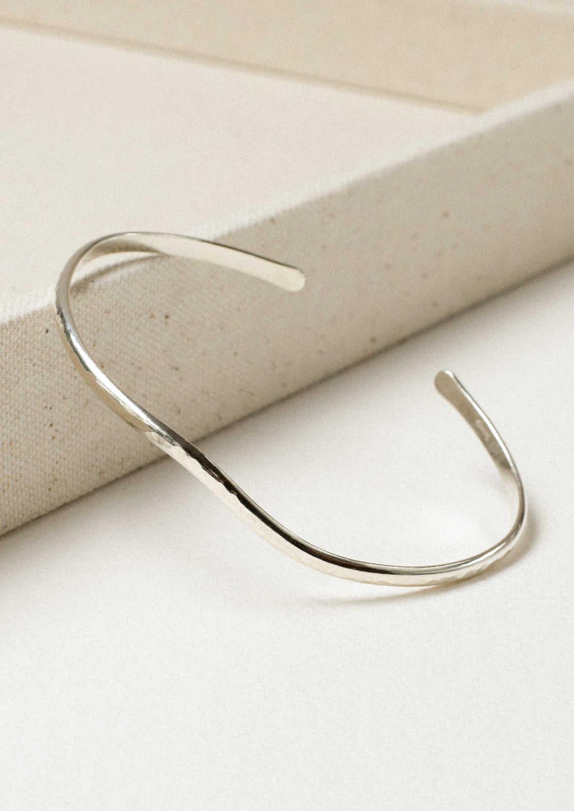 A minimal silver cuff bracelet with a wave-like design leaning on the edge of a canvas box.