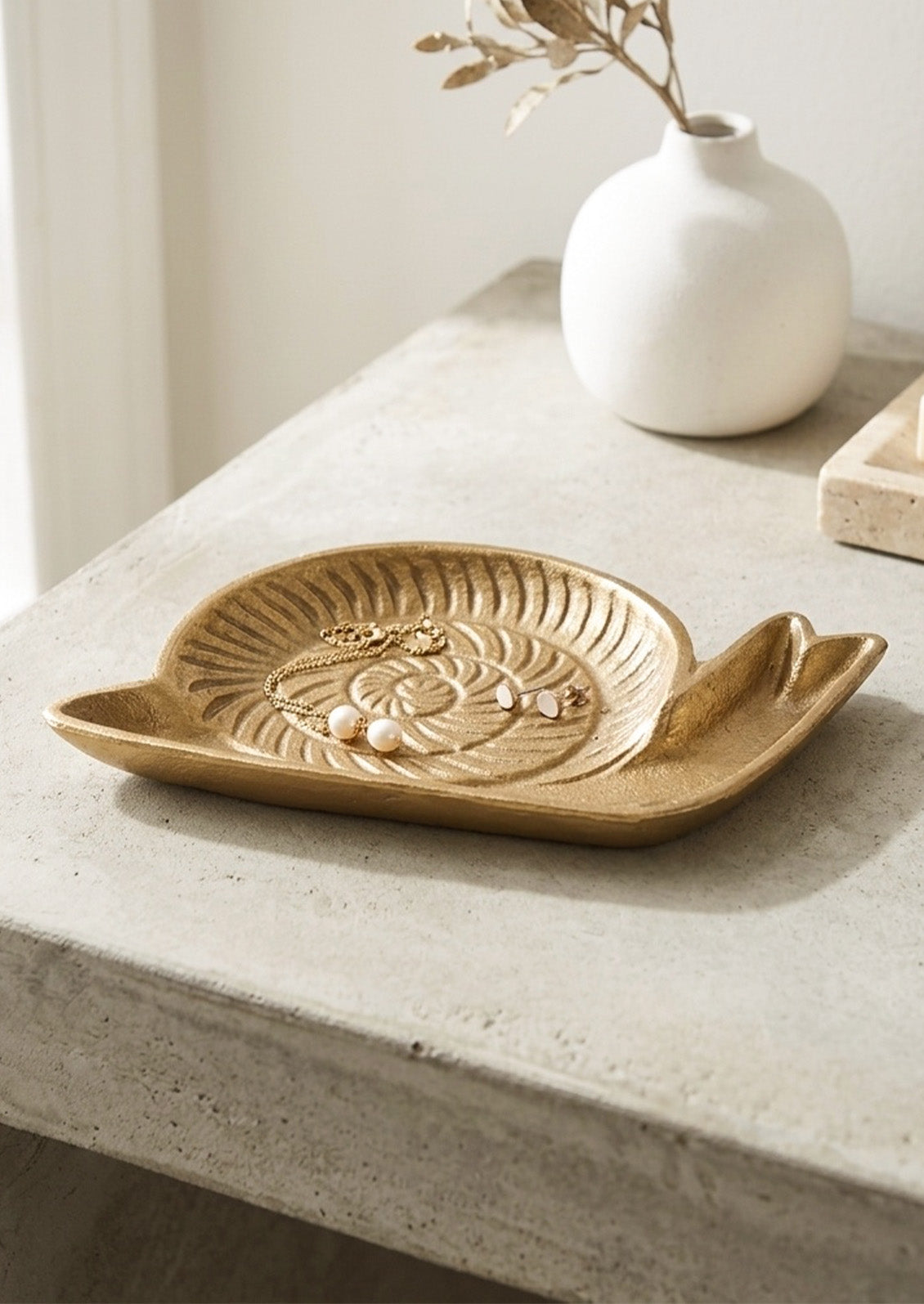 A handcrafted cast iron catchall tray in the shape of s snail in a gold finish holding jewelry and sitting on a stone table.