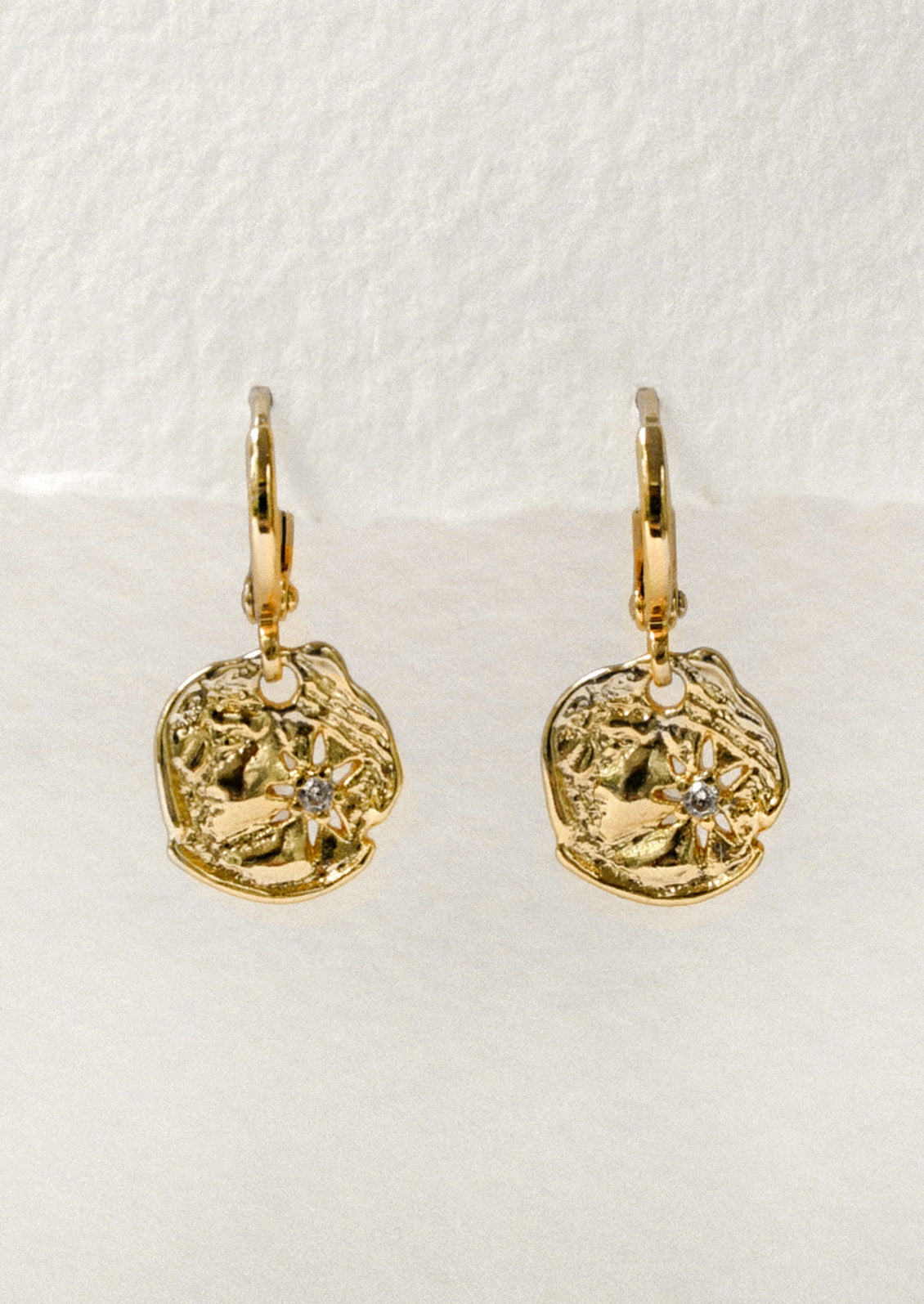 A pair of gold huggie hoop earrings with dangling disc charms that have a starburst design and a cubic zirconia in the center.