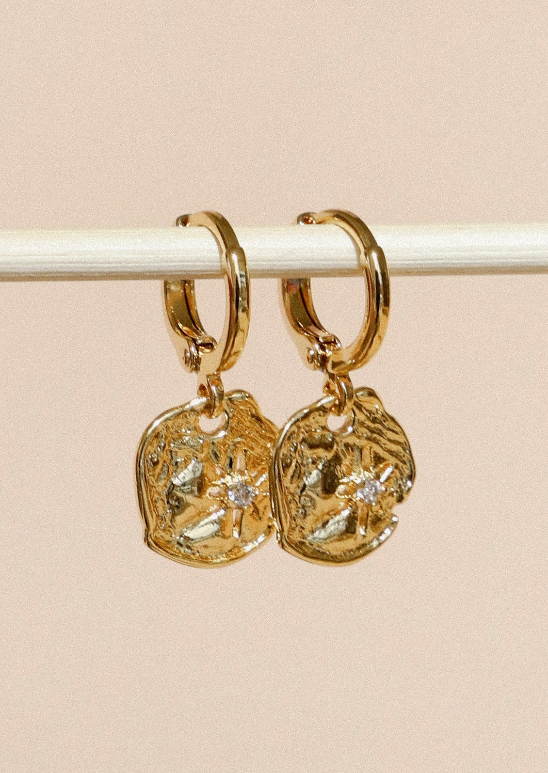 A pair of gold huggie hoop earrings with dangling disc charms that have a starburst design and a cubic zirconia in the center, hanging from a wooden stick in front of a beige background.