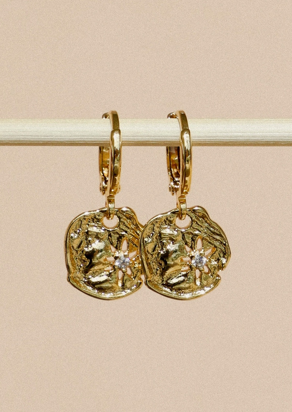 A pair of gold huggie hoop earrings with dangling disc charms that have a starburst design and a cubic zirconia in the center, hanging from a wooden stick in front of a beige background.