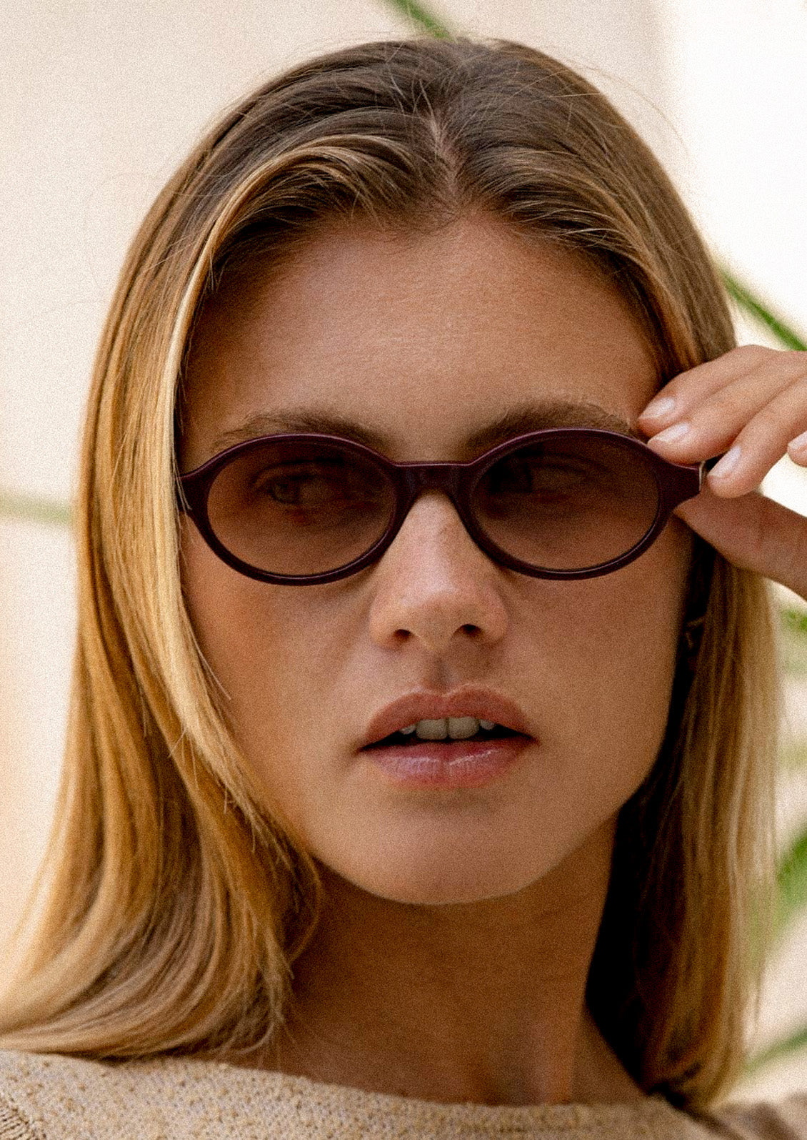 A woman wearing a pair of round oval sunglasses with a burgundy frame and light brown tinted lenses.