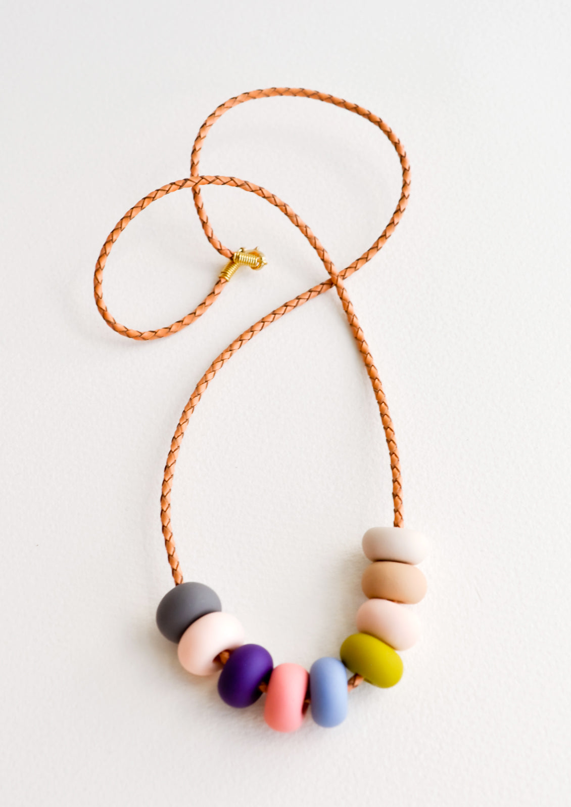 Woven leather cord necklace with gold clasp and rounded clay beads tans, pinks, purple, blue, gray, and chartreuse.