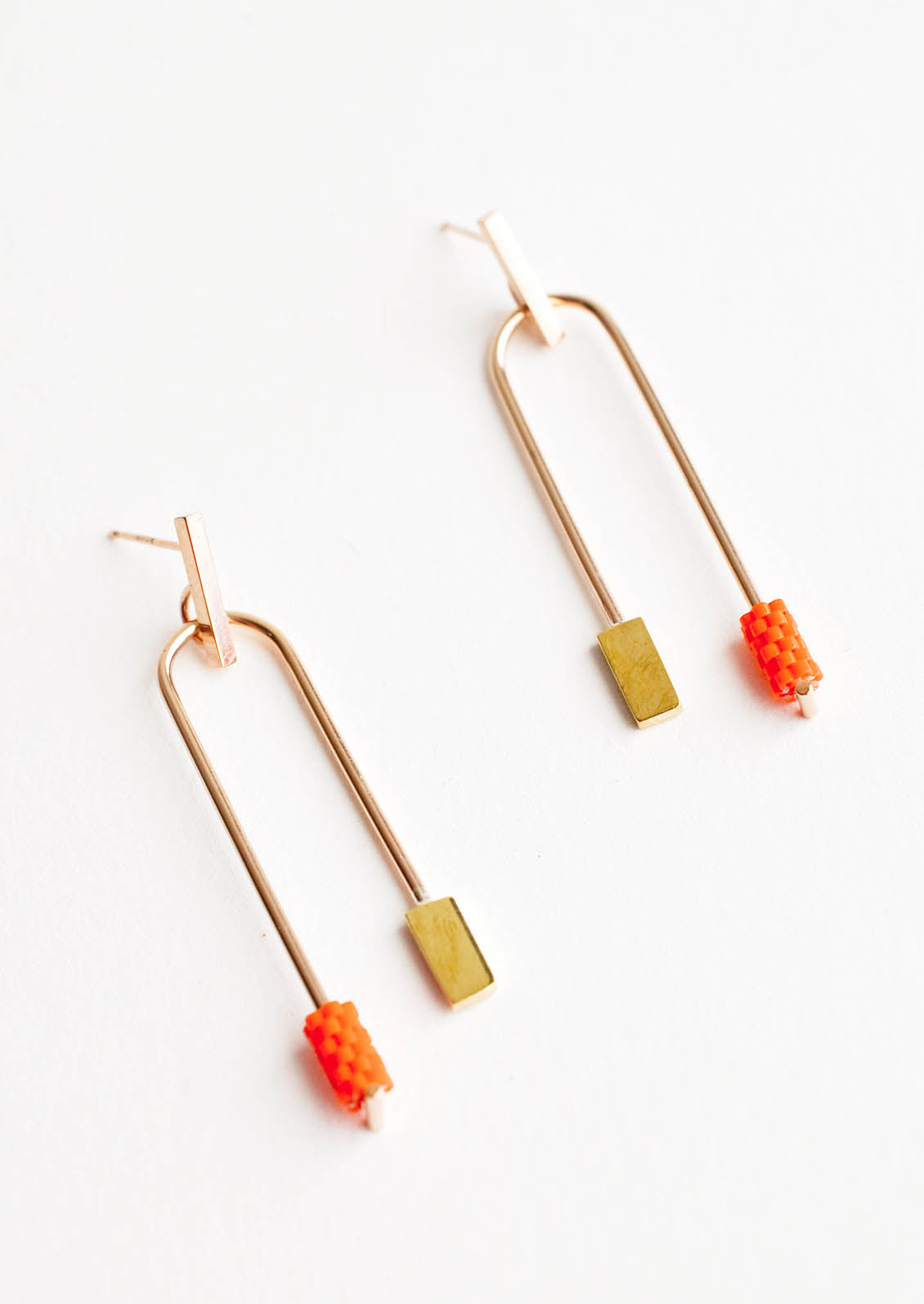 Post back gold earrings with curved arc featuring one orange beaded end and one square gold end.