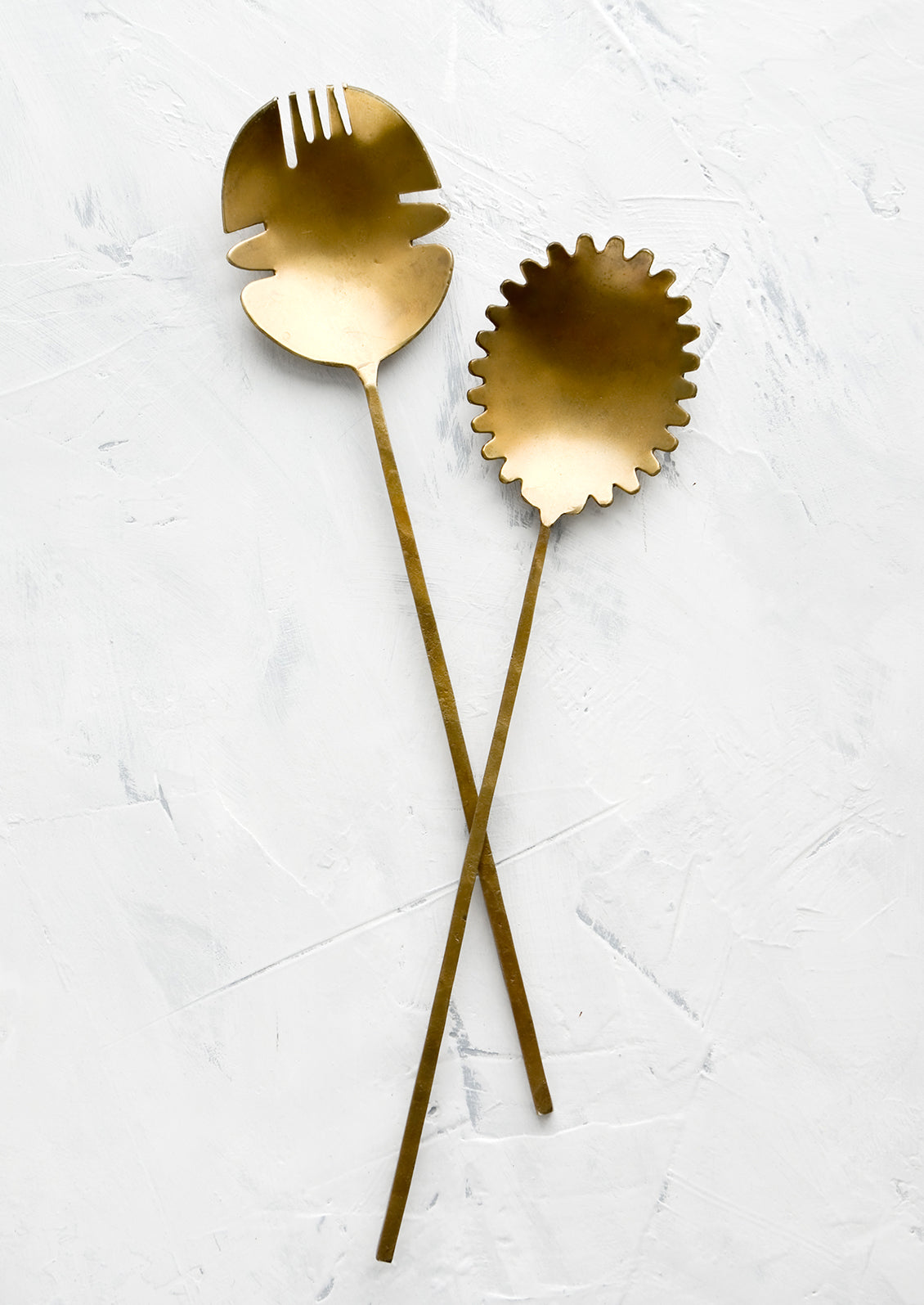 A pair of salad servers made from antiqued brass, shaped in a prehistoric-inspired design with slender handles.